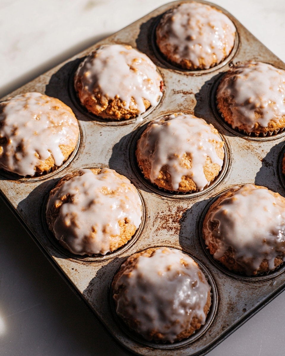 The image shows a metal muffin tray holding seven muffins, each topped with a shiny white glaze that covers the bumpy textured surface fully. The muffins are golden brown and look soft with some small uneven bits peeking through the smooth glaze. The muffin tray has a worn look with light scratches and spots, placed on a white marbled surface. Bright, natural light casts soft shadows across the muffins and tray, giving the photo depth and warmth. photo taken with an iphone --ar 4:5 --v 7