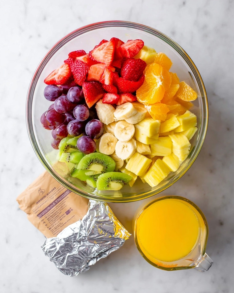 A clear glass bowl on a white marbled surface holds a colorful mix of six groups of fruit arranged in sections. Starting from the top left, there are bright red sliced strawberries, next to small yellow pineapple chunks, then orange segments on the right. Below the orange, green kiwi slices with black seeds form the fifth section, next to a cluster of red grapes, some whole and some cut in half showing the green inside. In the center of the bowl, pale yellow banana slices are stacked on top. Below the bowl on the surface are a silver wrapped block of cream cheese, a brown paper bag, and a clear glass measuring cup filled with bright orange liquid. Photo taken with an iphone --ar 4:5 --v 7